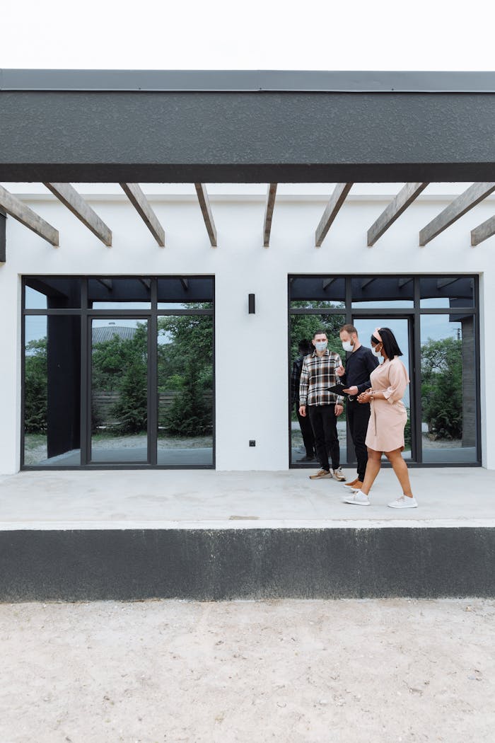 gallery-2 Three real estate agents wearing masks showcase a sleek, modern home exterior to clients.